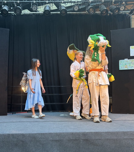 KI generiert: Das Bild zeigt eine Bühnenaufführung, bei der ein Kind im blauen Kleid neben zwei anderen Kindern steht, die einen grünen und gelben Drachenkostümkopf tragen. Die Szene scheint Teil einer Schul- oder Gemeinschaftsvorstellung zu sein.