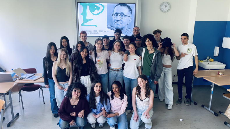 KI generiert: Eine Gruppe von Schülern steht in einem Klassenraum vor einem Bildschirm, der ein Porträt und Informationen über Bertolt Brecht zeigt. Es handelt sich um ein Klassenfoto im Rahmen einer Präsentation oder eines Projekts über den Schriftsteller.