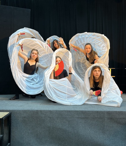 KI generiert: Das Bild zeigt fünf Frauen in schwarzen Trikots, die innerhalb großer, spiralförmiger, weißer Stoffstrukturen posieren und rote Fächer in den Händen halten. Sie stehen auf einer Bühne vor einem schwarzen Hintergrund.