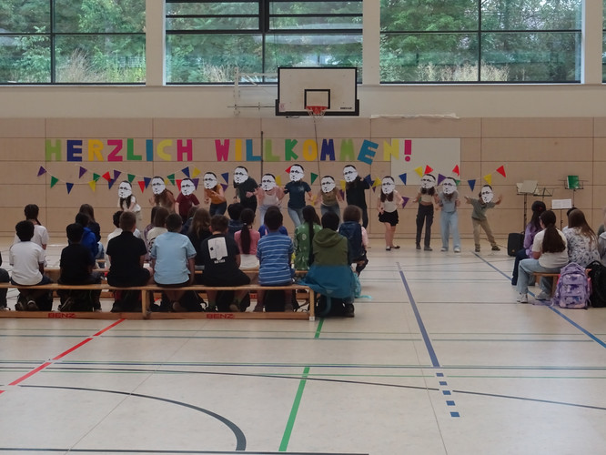 KI generiert: Das Bild zeigt eine Gruppe von Kindern, die auf einer Bühne stehen und große Gesichtsmasken tragen, während sie von einem Publikum aus anderen Kindern in einer Turnhalle betrachtet werden. Im Hintergrund hängt ein buntes Banner mit der Aufschrift "Herzlich Willkommen".