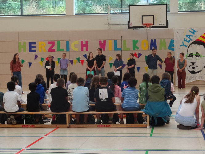 KI generiert: Das Bild zeigt eine Gruppe von Menschen in einer Turnhalle, die eine Begrüßungsveranstaltung mit dem Schriftzug "Herzlich Willkommen" durchführt. Vorne sitzen Kinder auf Bänken und schauen nach vorne.