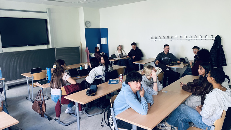 KI generiert: Das Bild zeigt eine Gruppe von Schülern, die in einem Klassenzimmer an Tischen sitzen. Sie unterhalten sich miteinander, während Tablets und andere Lernmaterialien auf den Tischen liegen.