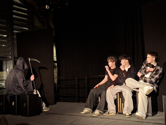 KI generiert: Auf dem Bild ist eine Bühnenszene zu sehen, in der eine Person in einem dunklen Kapuzenumhang mit einer Sense drei sitzenden jungen Männern gegenübersteht. Die Szene wirkt wie eine Theaterdarstellung mit mysteriösem oder dramatischem Thema.