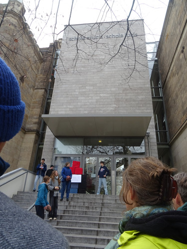 KI generiert: Das Bild zeigt eine Gruppe von Menschen, die vor einem Gebäude mit der Aufschrift "Schola" stehen. Einige Personen unterhalten sich auf der Treppe vor dem Eingang.