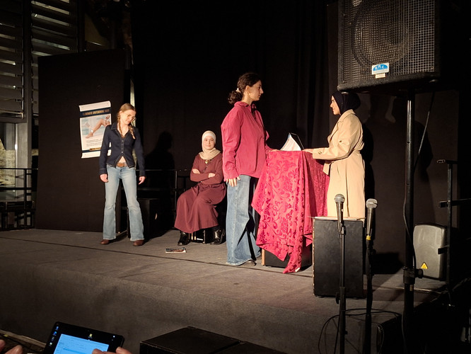 KI generiert: Das Bild zeigt eine Theateraufführung mit vier Personen auf einer Bühne, wobei eine davon sitzt und die anderen diskutieren oder interagieren. Im Vordergrund steht ein Tisch mit einem roten Tuch, vor dem zwei Personen miteinander sprechen.