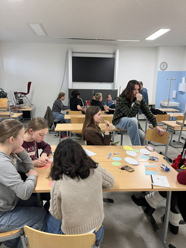 KI generiert: Das Bild zeigt eine Gruppe von Schülern in einem Klassenzimmer, die zusammen an einem Tisch sitzen und mit Scheren und buntem Papier arbeiten. Im Hintergrund unterhalten sich weitere Schüler an anderen Tischen.