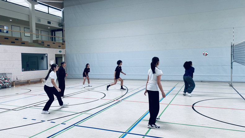 KI generiert: Das Bild zeigt eine Gruppe junger Frauen in einer Sporthalle, die Volleyball spielen. Eine von ihnen ist dabei, den Ball am Netz zurückzuschlagen.