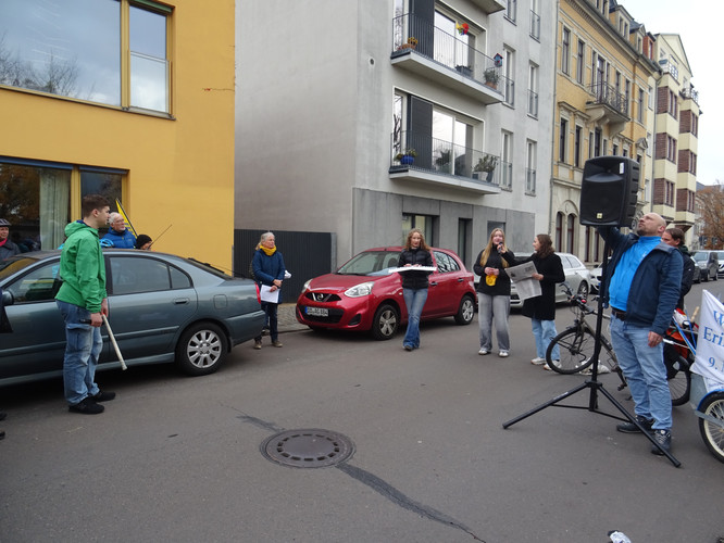 KI generiert: Auf dem Bild sind mehrere Personen auf einer Straße versammelt. Sie stehen um einen Lautsprecher, was auf eine kleine Versammlung oder Kundgebung hinweisen könnte.