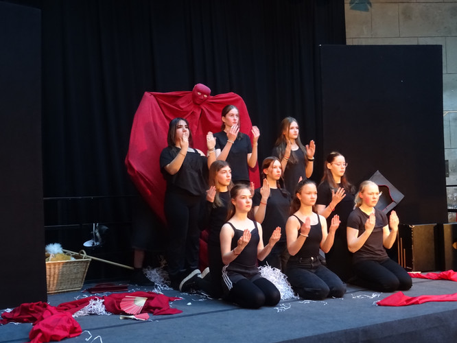 KI generiert: Auf dem Bild ist eine Gruppe von Personen zu sehen, die auf einer Bühne performt und schwarze Kleidung trägt; im Hintergrund gibt es eine Figur mit einem roten Umhang. Die Szene wirkt inszeniert und dramatisch, möglicherweise Teil einer Theateraufführung.