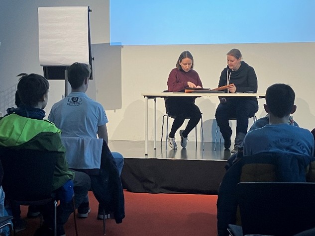 KI generiert: Das Bild zeigt zwei Personen, die an einem Tisch auf einer Bühne sitzen und ein Buch betrachten, während mehrere Menschen im Publikum sitzen und zuschauen. Im Hintergrund steht ein Flipchart.