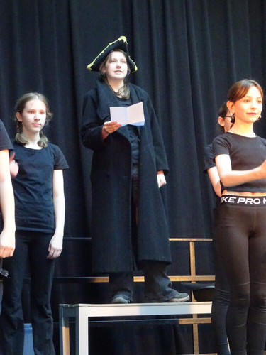 KI generiert: Das Bild zeigt eine Gruppe junger Menschen auf einer Bühne, wobei eine Person im Vordergrund im schwarzen Mantel und mit einem Dreieckshut eine Rolle spielt. Diese Person hält ein Blatt Papier und scheint, als ob sie etwas vorträgt oder vorliest.