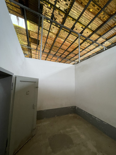 KI generiert: Das Bild zeigt einen leeren Raum mit einem offenen Gitterdach, durch das Sonnenlicht scheint. Die Wände sind weiß gestrichen, und eine geöffnete Metalltür ist links im Bild sichtbar.