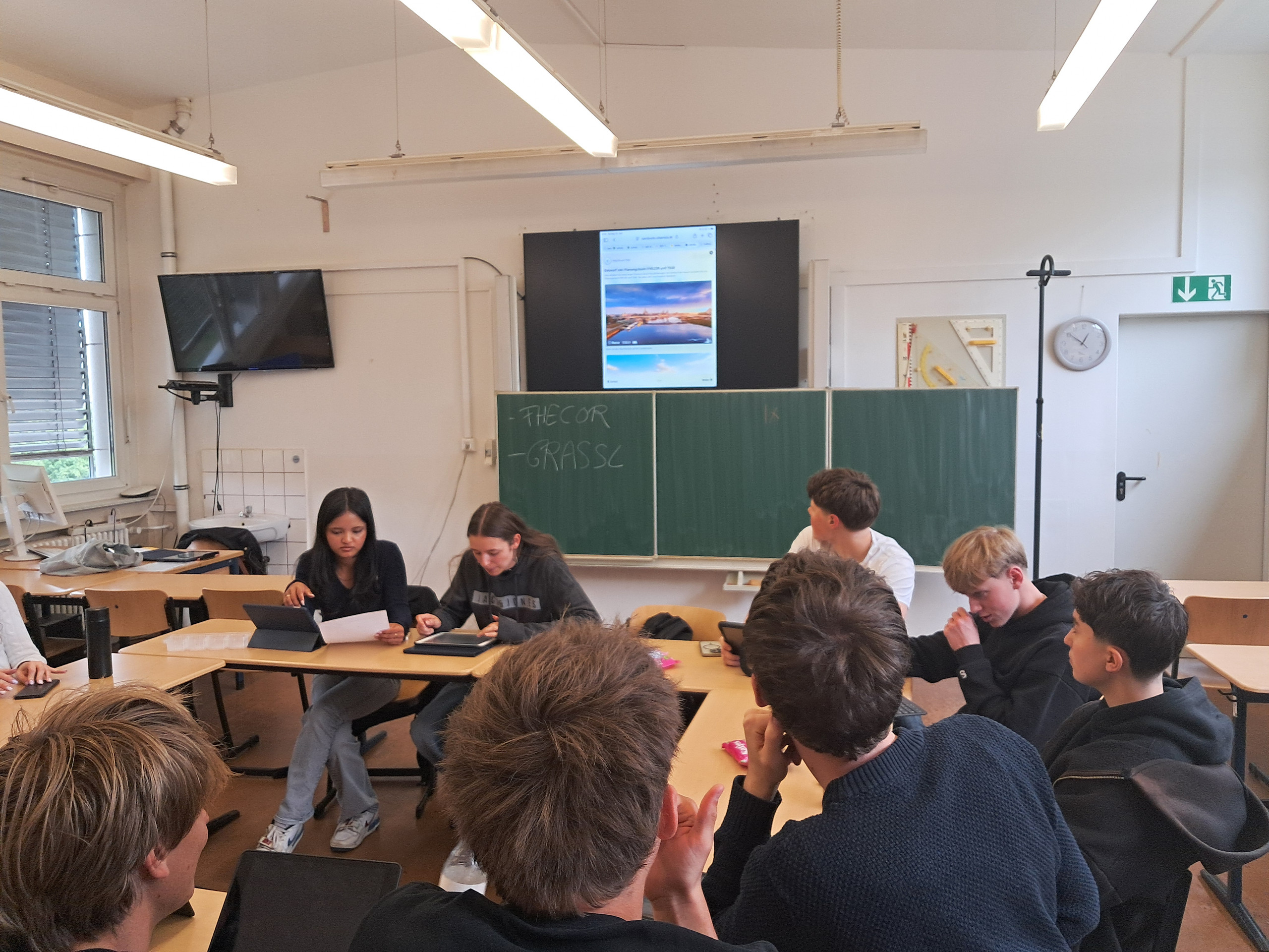 KI generiert: Eine Gruppe von Schülerinnen und Schülern sitzt in einem Klassenzimmer und diskutiert, während auf einem großen Bildschirm ein Bild angezeigt wird. An der Tafel stehen die Wörter "THEO R" und "GRASS C".