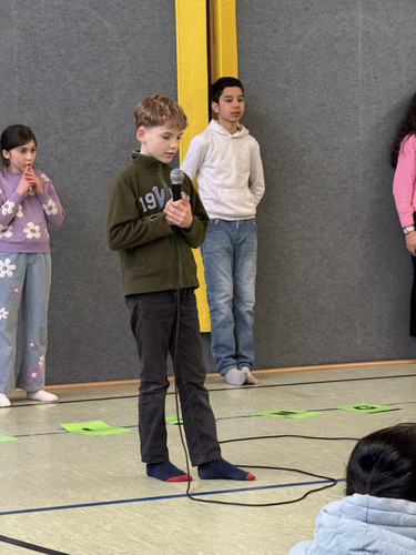 KI generiert: Ein Kind steht mit einem Mikrofon in der Hand vor einer Gruppe von Menschen in einer Halle. Es sieht so aus, als ob es eine Präsentation oder Rede hält, während andere Kinder im Hintergrund stehen.