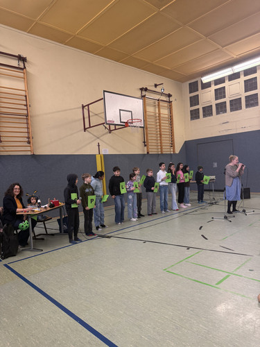 KI generiert: Das Bild zeigt eine Gruppe von Kindern, die in einer Turnhalle stehen und Buchstaben auf grünen Schildern halten, während eine Frau vor einem Mikrofon spricht. Die Kinder stehen in einer Reihe vor einer Tischgruppe, an der zwei Personen sitzen.
