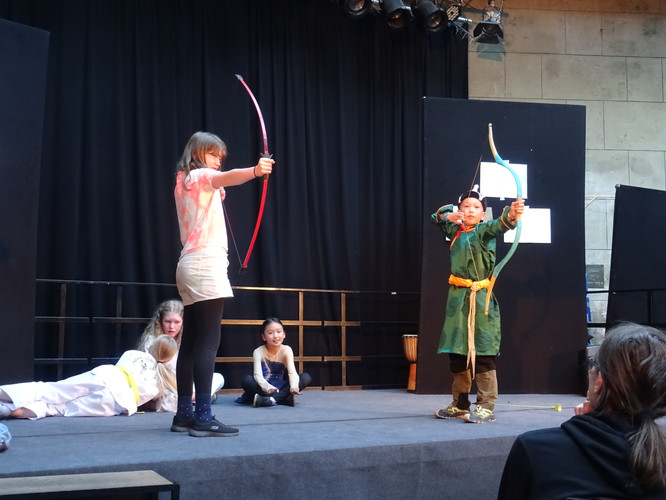 KI generiert: Auf dem Bild stehen zwei Kinder auf einer Bühne und halten jeweils einen Bogen, als ob sie schießen würden. Im Hintergrund sitzen weitere Kinder, die zuschauen.