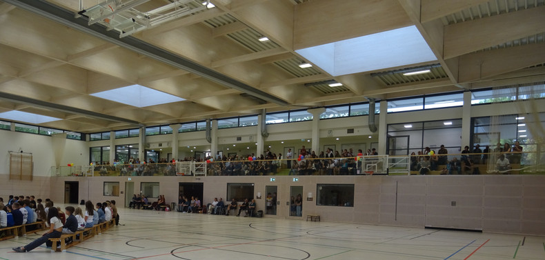 KI generiert: Das Bild zeigt eine Sporthalle mit Menschen, die auf Bänken sitzen und auf einer Galerie stehen. Der Fokus liegt auf den Zuschauern, die offenbar einer Veranstaltung beiwohnen.