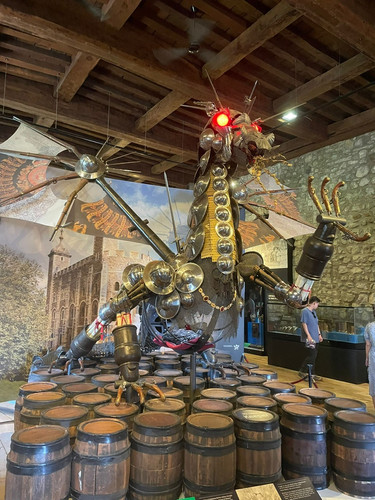 KI generiert: Das Bild zeigt eine große Drachenskulptur, die aus verschiedenen Metallobjekten und Schilden zusammengesetzt ist. Der Drache hat leuchtend rote Augen und ist von mehreren Fässern umgeben.