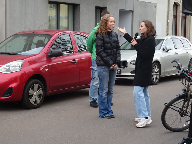 KI generiert: Im Bild unterhalten sich zwei Frauen auf einer Straße, während eine ein Mikrofon hält. Im Hintergrund sind parkende Autos zu sehen.