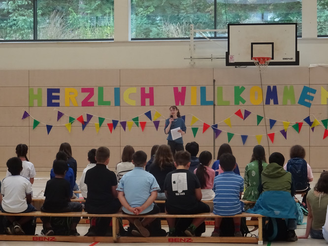 KI generiert: Das Bild zeigt eine Begrüßungsveranstaltung in einer Turnhalle mit einem "Herzlich Willkommen"-Banner im Hintergrund. Eine Person spricht zu einer Gruppe von sitzenden Kindern.