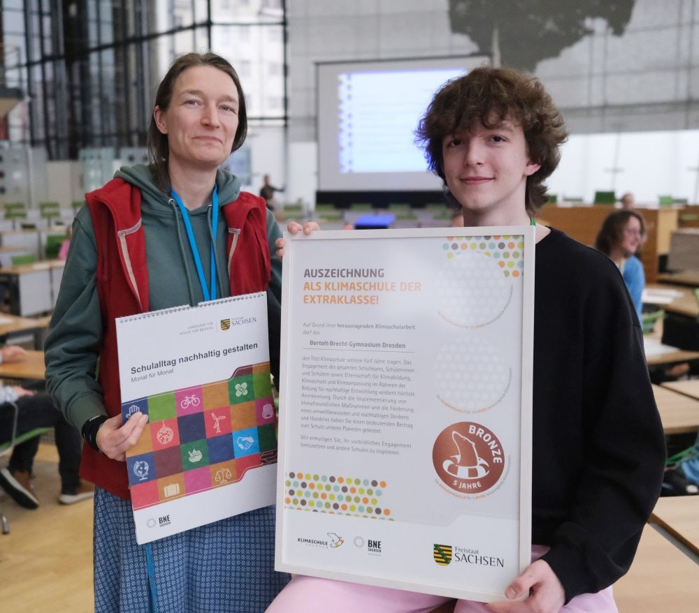 KI generiert: Das Bild zeigt zwei Personen, die ein Zertifikat und ein Poster mit der Aufschrift "Auszeichnung als Klimaschule der Extraklasse" in einem Raum mit mehreren Tischen und Stühlen halten. Es handelt sich um eine Anerkennung für herausragende Klimaschutzarbeit.