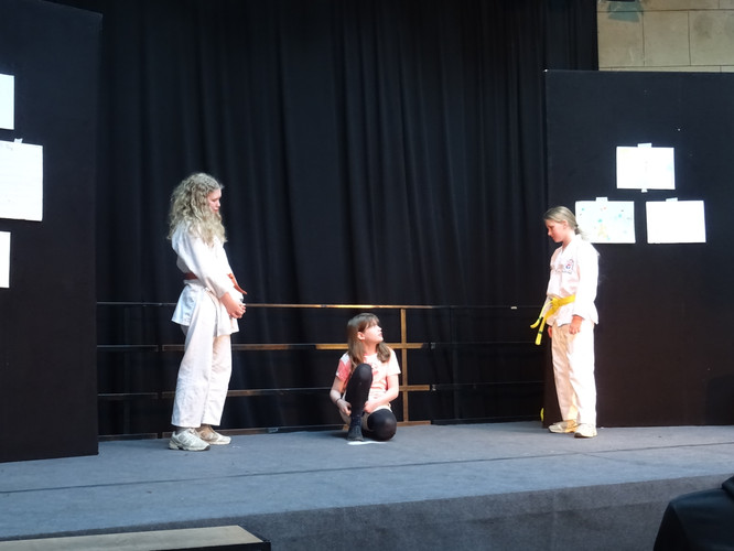 KI generiert: Das Bild zeigt eine Theateraufführung mit drei Kindern auf einer Bühne, von denen zwei weiß gekleidet sind und Gürtel tragen. Eines der Kinder sitzt, während die anderen beiden stehen und miteinander interagieren.