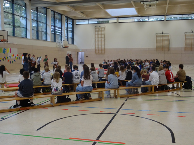 KI generiert: Das Bild zeigt eine Versammlung in einer Sporthalle, bei der eine Gruppe von Personen auf Bänken sitzt und eine kleinere Gruppe von Menschen vorne eine Präsentation oder Aufführung hält. Die Anordnung und Umgebung deuten auf eine schulische Veranstaltung hin.