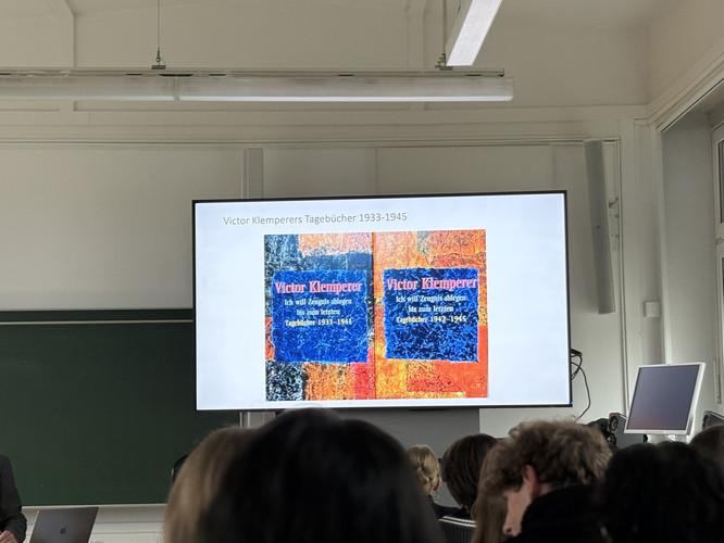 KI generiert: In dem Bild wird eine Präsentation mit Büchern von Victor Klemperer gezeigt, auf der ein Bildschirm seine Tagebücher von 1933-1945 präsentiert. Im Hintergrund befinden sich Teilnehmer in einem Raum mit Tafel und moderner Technik.