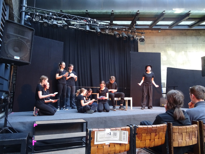 KI generiert: Das Bild zeigt eine Gruppe von jungen Leuten auf einer Bühne, die anscheinend an einer Theaterprobe oder -aufführung teilnehmen. Sie tragen schwarzes Bühnenoutfit und ein Mädchen steht im Vordergrund, während die anderen sitzen oder stehen.