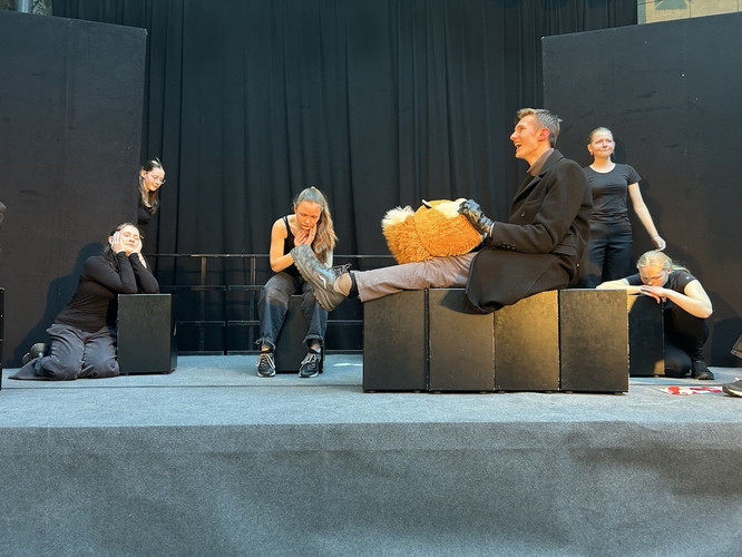 KI generiert: Das Bild zeigt eine Theaterprobe, bei der ein Darsteller mit einem großen Plüschspielzeug auf der Bühne sitzt, umgeben von anderen Schauspielern in schwarzen Outfits. Sie scheinen verschiedene Szenen zu proben oder humorvolle Interaktionen darzustellen.