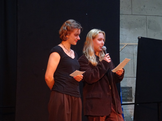 KI generiert: Zwei Personen stehen auf einer Bühne und halten jeweils ein Blatt Papier; eine der Personen spricht in ein Mikrofon. Der Hintergrund ist dunkel, was den Fokus auf die sprechenden Personen legt.