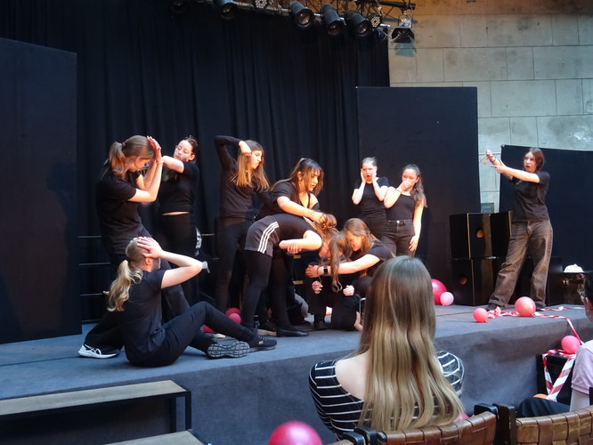 KI generiert: Das Bild zeigt eine Gruppe von Personen auf einer Bühne, die sich in einer Art theatralischer Performance befinden. Einige der Personen nehmen expressive Körperhaltungen ein, während sie scheinbar eine Szene darstellen.