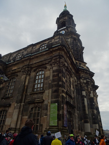 KI generiert: Das Bild zeigt eine beeindruckende, historische Kirche mit einem Turm, bei der zahlreiche Menschen davor Versammlungen oder eine Demonstration abhalten. Einige der Teilnehmer halten Schilder hoch.