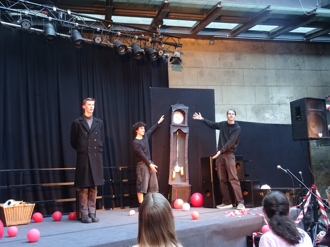 KI generiert: Das Bild zeigt eine Theaterszene mit drei Personen auf einer Bühne, die sich um eine hohe Standuhr versammeln. Im Vordergrund sind rote Luftballons und einige Zuschauer zu sehen.