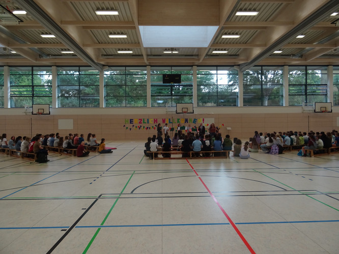 KI generiert: Das Bild zeigt eine Schulveranstaltung in einer Turnhalle, bei der zahlreiche Menschen auf Bänken sitzen und einer Präsentation auf einer Bühne mit einem "Herzlich Willkommen"-Banner zuschauen. Die Atmosphäre wirkt freundlich und einladend.